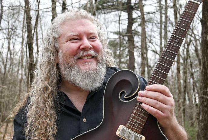 Joe Jencks holding his guitar, smiling in a forest.