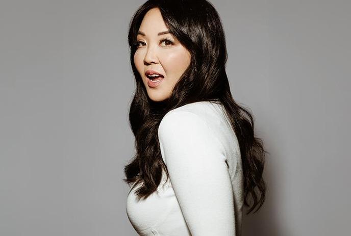 A lady with long, dark hair wearing a white long-sleeve shirt is standing sideways against a plain grey background.