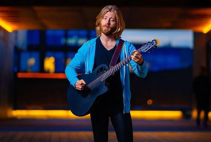 A person with a blurred face is playing a guitar in an urban setting with warm lighting and a blue sky in the background.