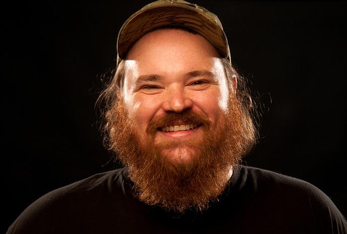 A bearded man wearing a cap smile in the foreground, with three smaller portraits of people beside him in dark background.