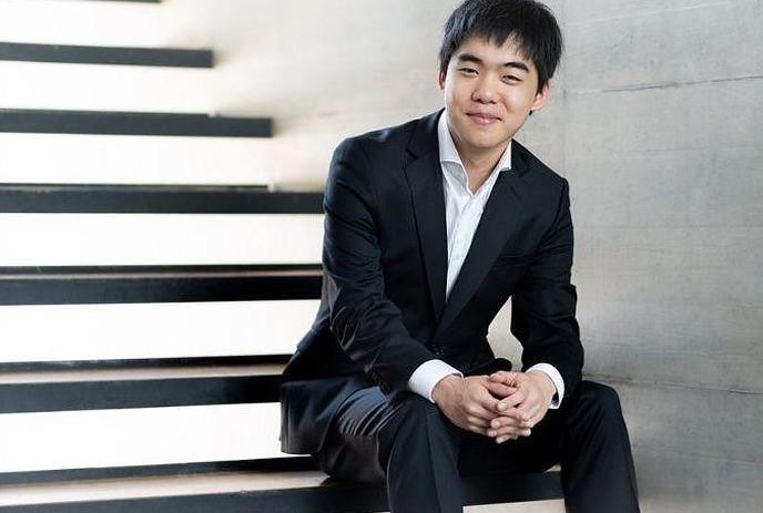 A person smiling, sitting on a set of white stairs. The individual is wearing a black suit .