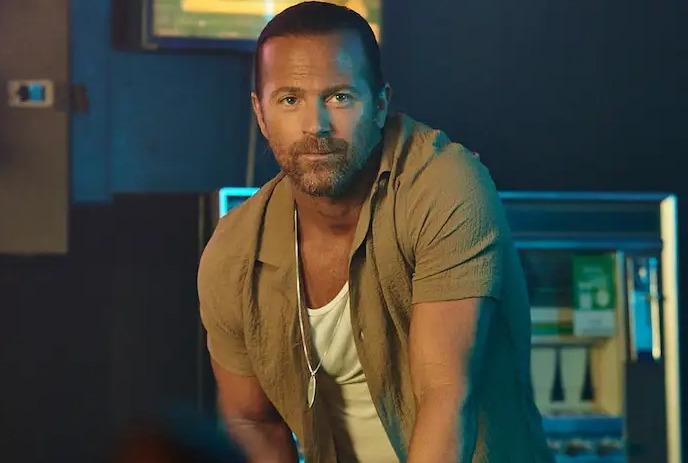 Kip Moore leaning forward in a dimly lit setting, wearing a tan shirt over a white tank top with a serious expression.