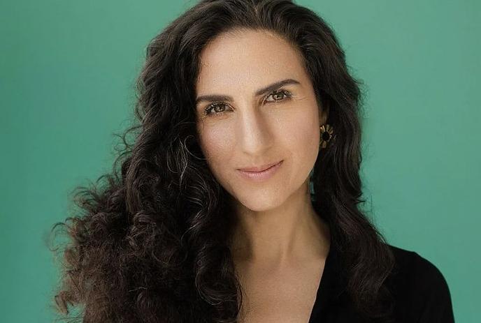 A woman with curly hair and smiling face stands against a teal background posing for the picture.