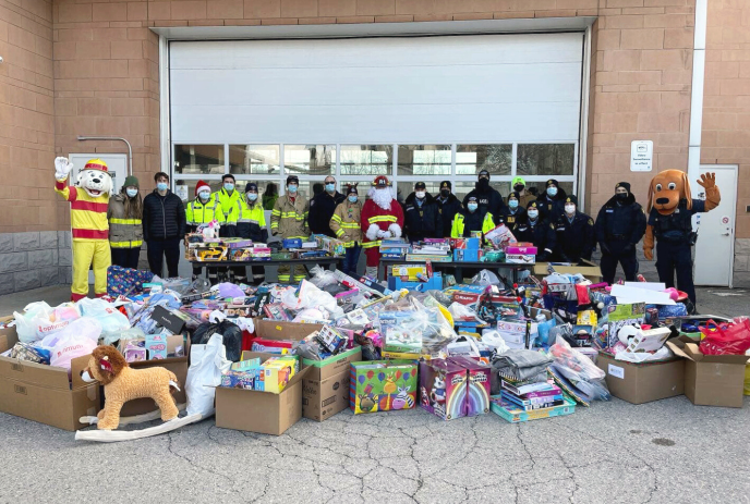 A group of people, including some in costumes and uniforms, stand behind tables filled with toys and boxes.