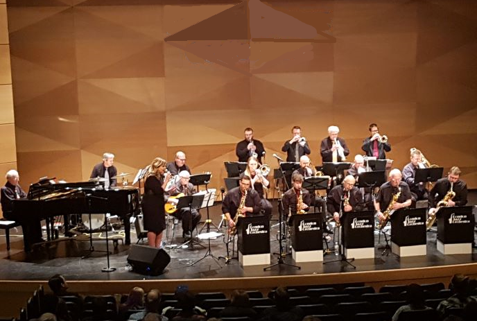 A jazz band performs on stage with various instruments including saxophones, trumpets, trombones, a piano, and a guitar.