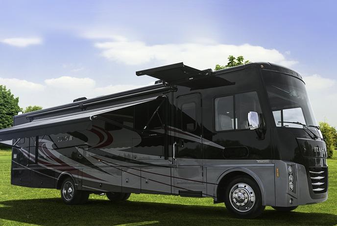 A white RV driving through a lush green mountainous landscape with towering rocky peaks in the background.