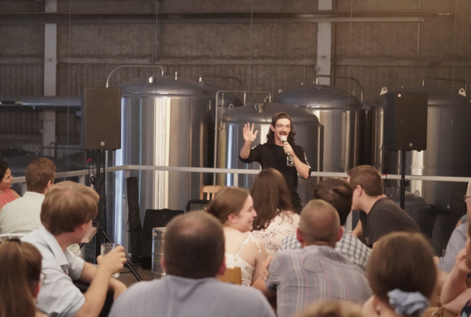 A  male comedian is gesturing while speaking to an audience in a room with large brewing tanks.