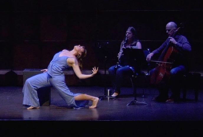 A dancer in a blue outfit performs a dramatic pose on stage, with two musicians playing instruments in the background.