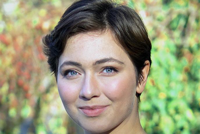 A female with short, dark hair is standing in front of a blurred, colorful background featuring green and red hues.