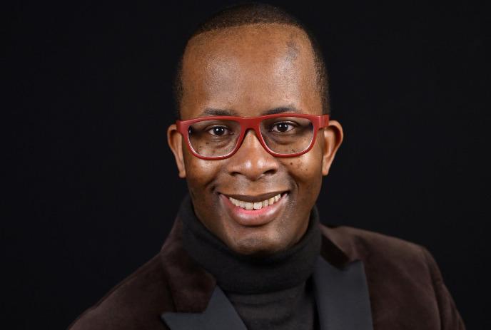 A person wearing red glasses and smiling warmly dressed in a dark turtleneck and a brown jacket.