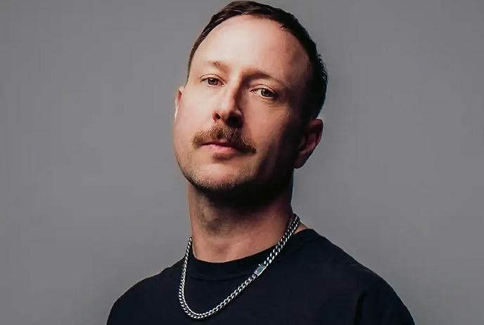 Marten Hørger wearing a black coat, standing against a white wall and smiling.