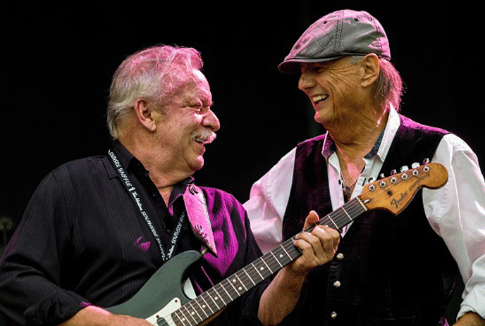 2 men performing on stage, looking at each other and smiling, one holding a guitar.