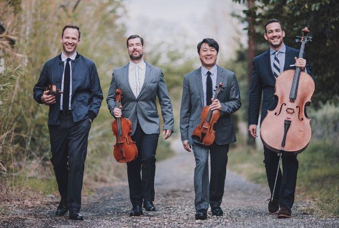 Four individuals in suits with string instruments on a path smiling for the picture. Greenery surrounds them.