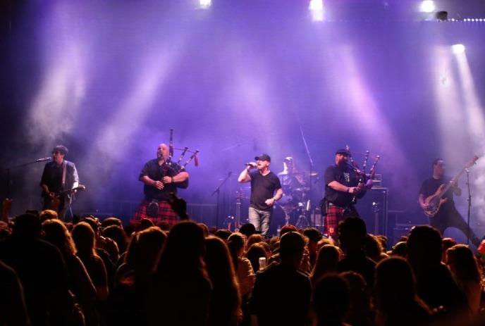 A band performs on stage with bagpipes, guitars, and drums, surrounded by a cheering crowd under purple lights.