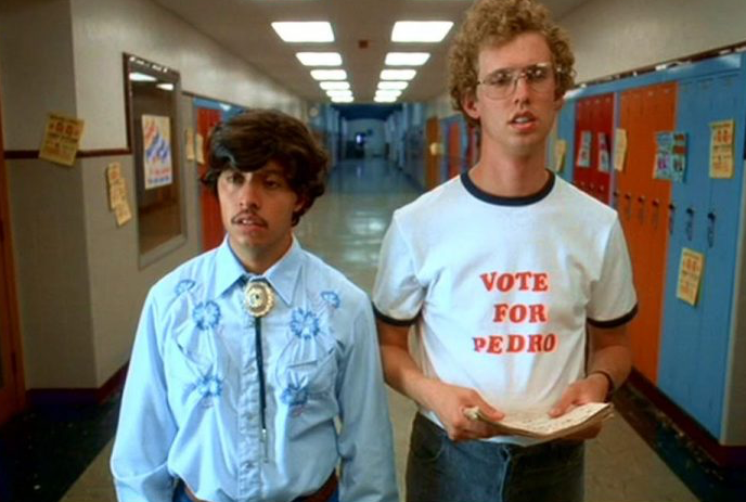 Two individuals in a school hallway, one with ‘Vote for Pedro’ shirt, faces obscured. Lockers and posters in the background.