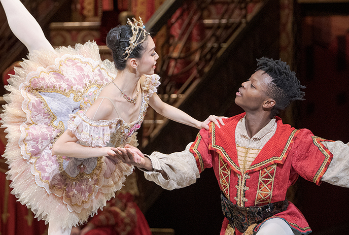 A ballerina in a pink and gold tutu and a male dancer in red and gold perform an elegant ballet duet on stage.