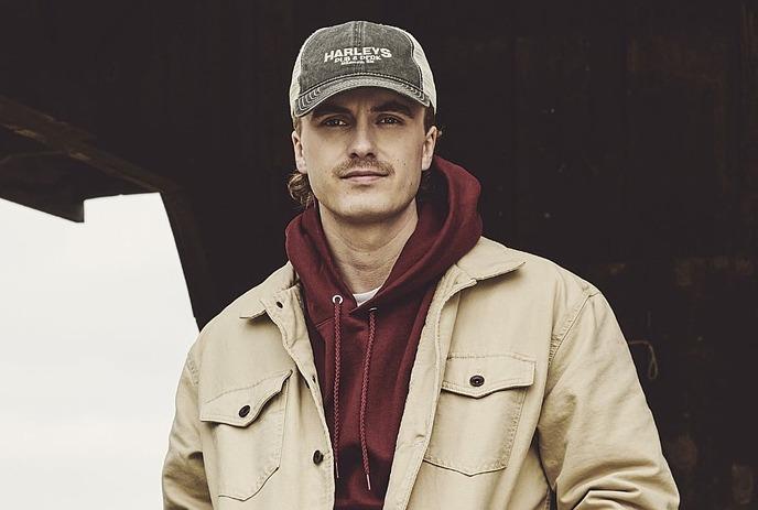 A portrait shot of Owen Riegling standing while wearing a beige jacket over a red hoodie and a cap.