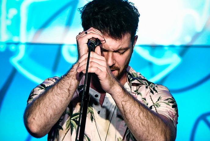 A man, with closed eyes, singing passionately into a microphone. He is wearing a floral shirt, with a dark background.