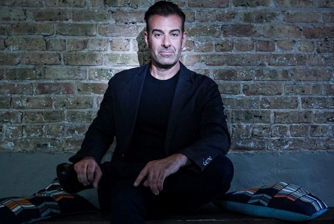 Paul Marinaro sitting down, posing for the camera in front of a brick wall in a room with dim lighting.