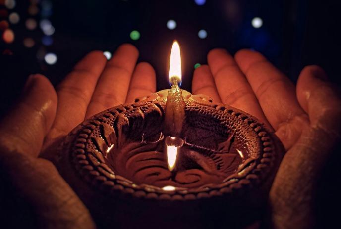 A pair of hands holding a lit candle with intricate designs, against a backdrop of colorful lights.