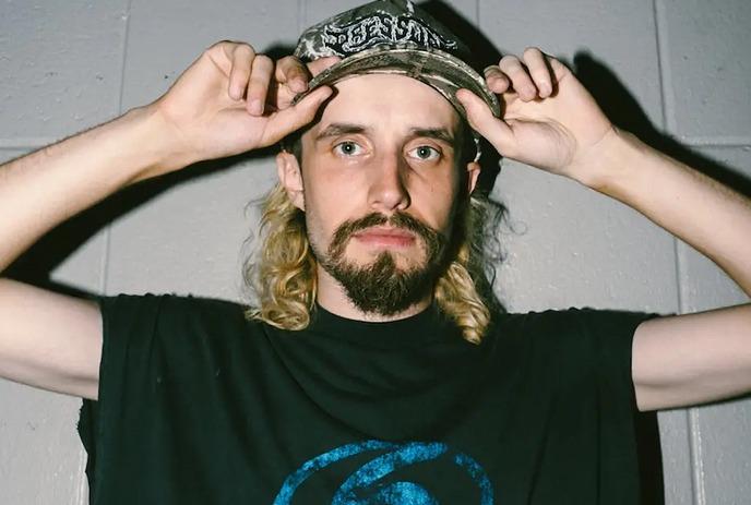 A person with long hair adjusting a patterned cap while wearing a black t-shirt with a blue circular logo.