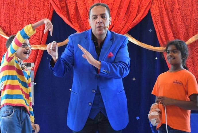 A person in a blue suit performs a magic trick with two children on stage, one in a striped shirt, other in an orange shirt.