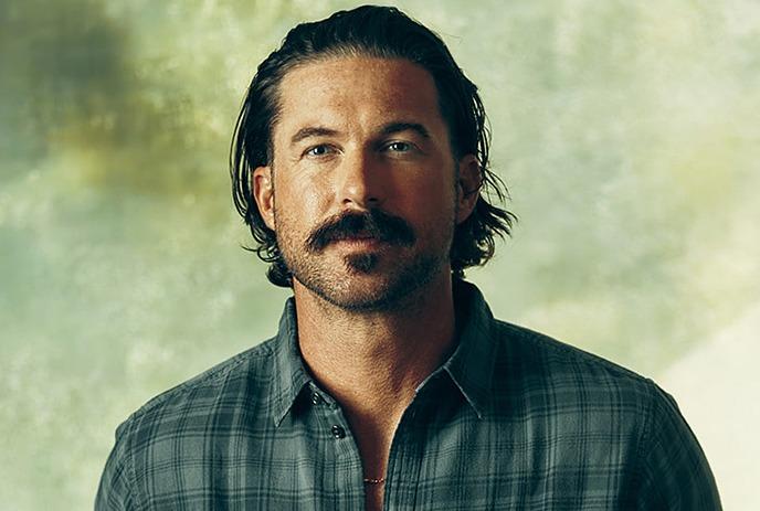 Riley Green looking at the camera wearing a plaid green shirt under a textured green and white background.