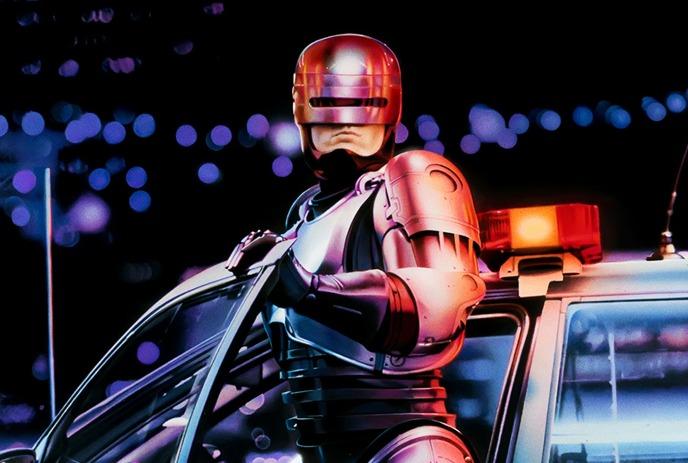 A futuristic, armored figure stands next to a police car with flashing lights in a night-time setting.