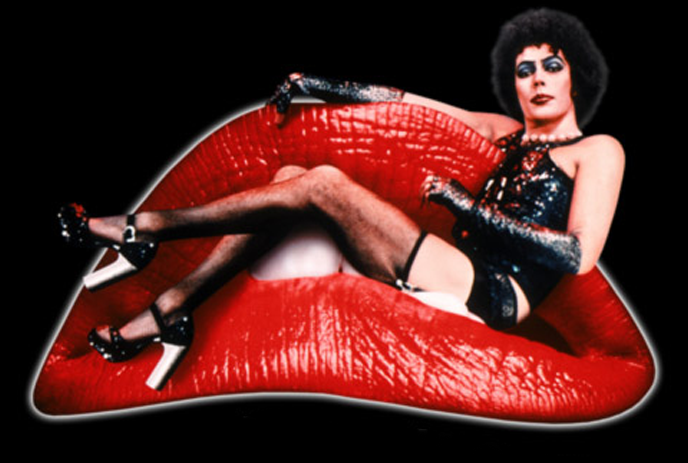 A person in glam rock attire in heels, is reclining confidently on a large red lip-shaped sofa against a black background.