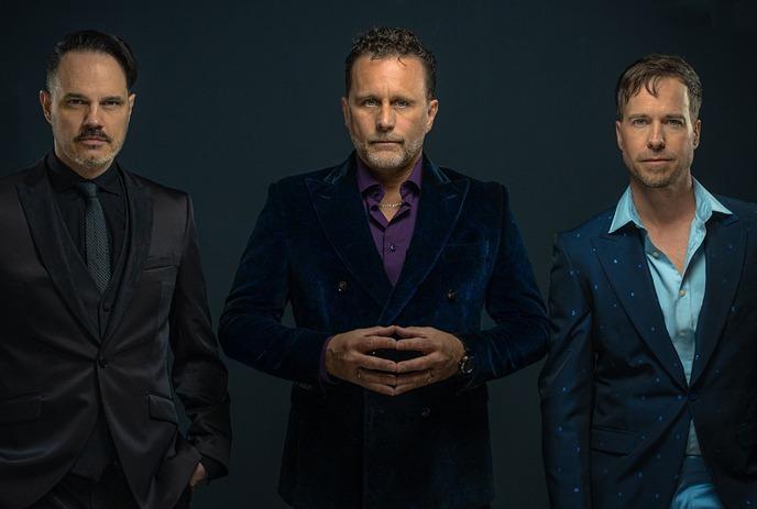 Three men in formal suits standing against a dark background, posing for the picture.