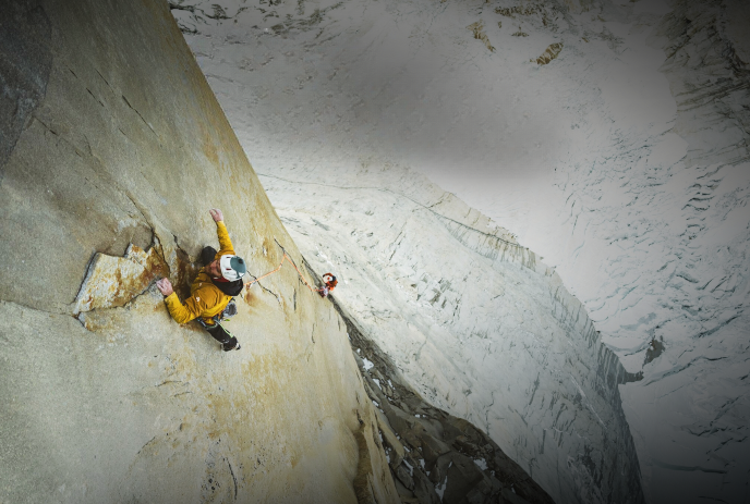 Two climbers, in yellow and red jackets, ascend a steep, rocky cliff face in a snowy, mountainous landscape.