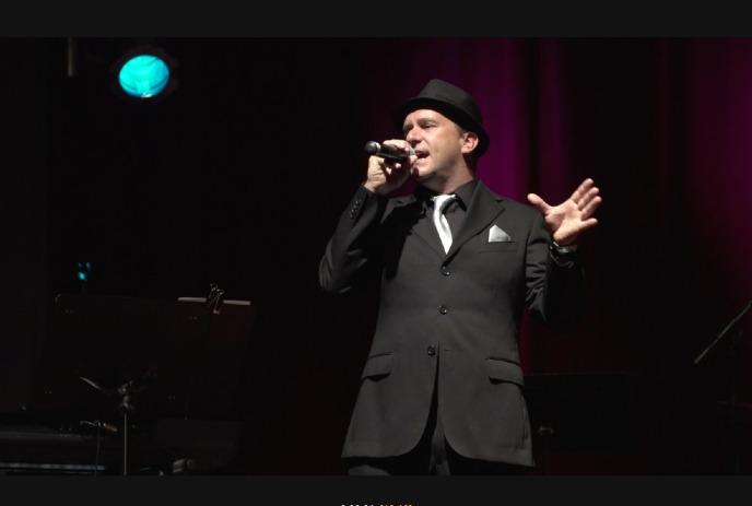 Scott Bollert performing as Frank Sinatra on stage with a microphone in his hand, wearing a suit.