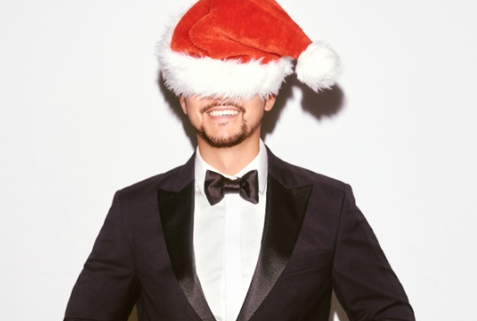 Person in a black suit, white shirt and Santa hat, smiling and standing against a plain background.