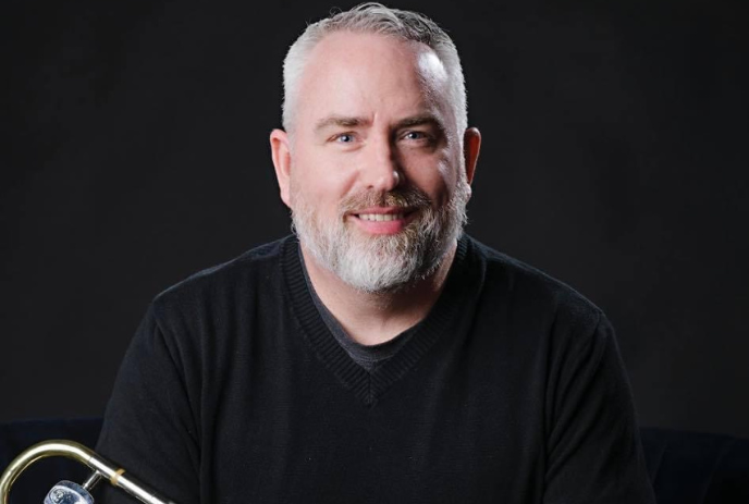 A man with short gray hair and a beard smile with a trombone, against a dark background.