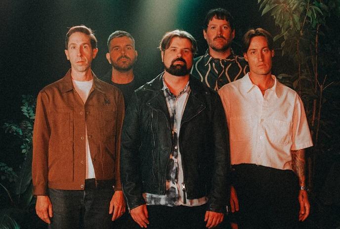 A group of five artists standing together in a dimly lit environment with green foliage in the background.