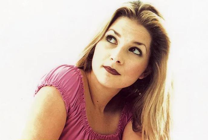 A woman in a pink top and black pants sitting on a chair, looking to her right, against a bright white background.