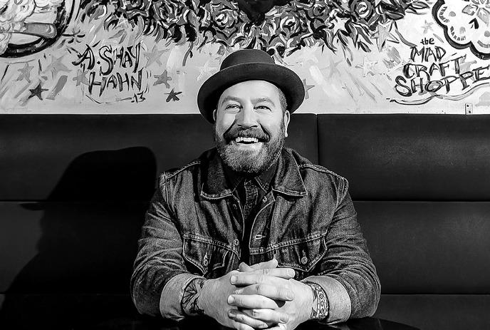 A black and white photo of Steve Marriner, sitting on a black couch, wearing a hat and smiling.