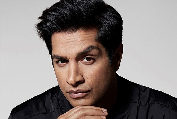 A close-up portrait of Sugar Sammy in black shirt in a serious face under a white background.