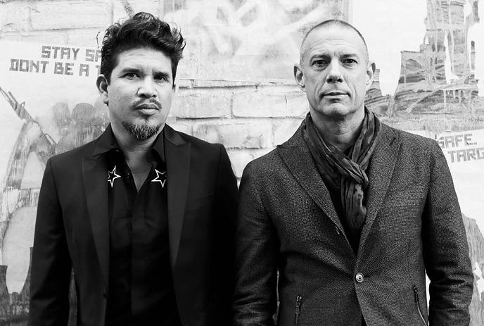 Two male artists in formal attire stand side by side against a graffiti-covered wall in the background,