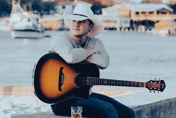 Thomas Mac sits with a guitar on his lap, leaning forward with his head down, next to a glass overlooking a waterfront.