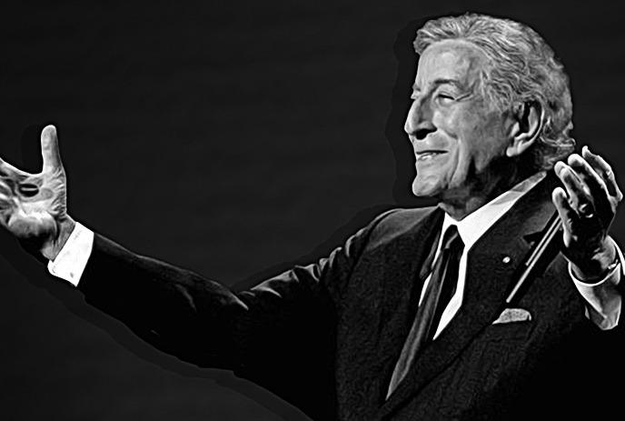 A black-and-white image of an older man in a suit, smiling and gesturing with both hands raised, against a dark background.