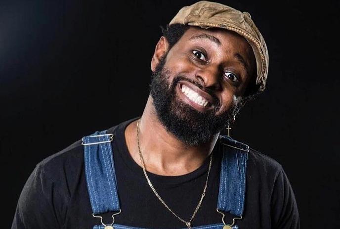 Jared Waters smiling and wearing a hat and a pair of overalls, against a black background.