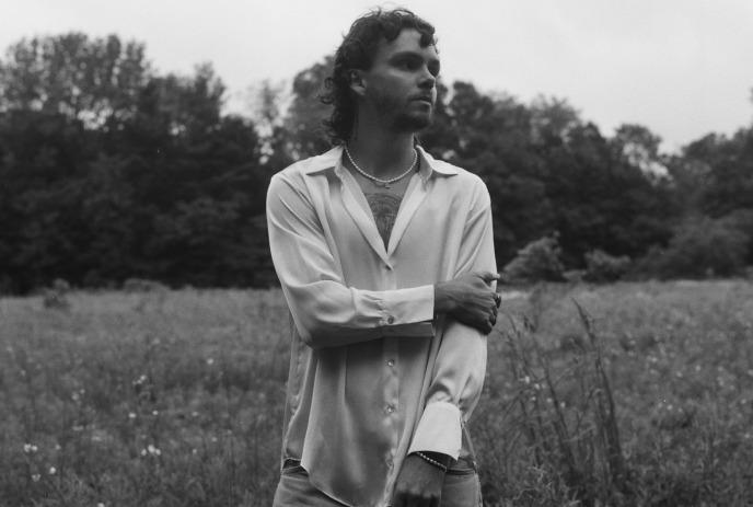 A man with curly hair, wearing a loose, open shirt, stands in a field, looking away thoughtfully.