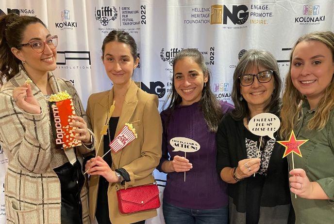 A group of five smiling women pose together at an event, holding playful movie-themed props, including popcorn and signs.