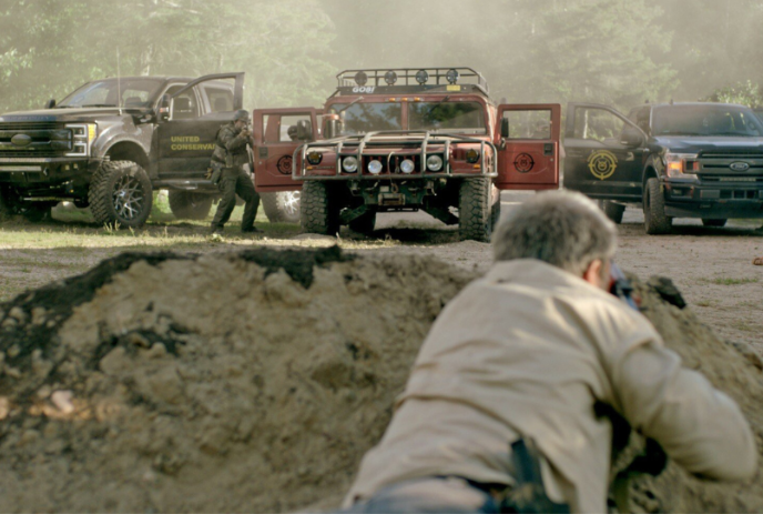 Person lying on their stomach aiming a firearm at a group of vehicles.