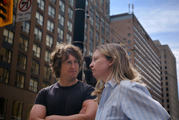 Two people standing in front of a large building looking concerned.