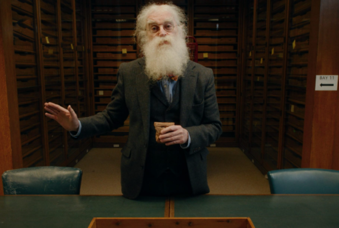 Older person with a white beard standing in a library in front of a game board.