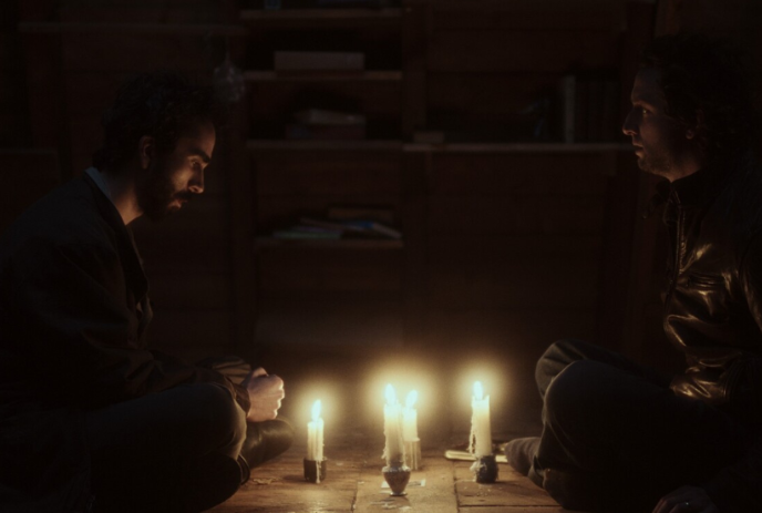 Two people sitting on the floor facing each other with a number of lit candles between them.