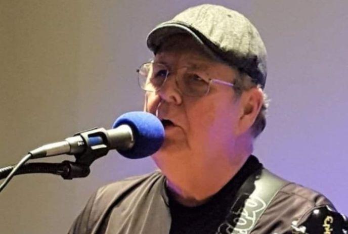 A portrait shot of Rick Watts performing on the stage on a microphone under a light background.