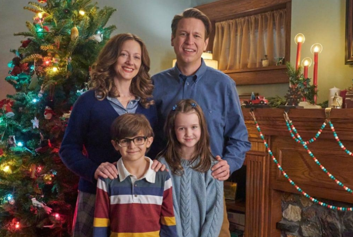 A mother, father and two kids stand in front of a Christmas tree and a fireplace.